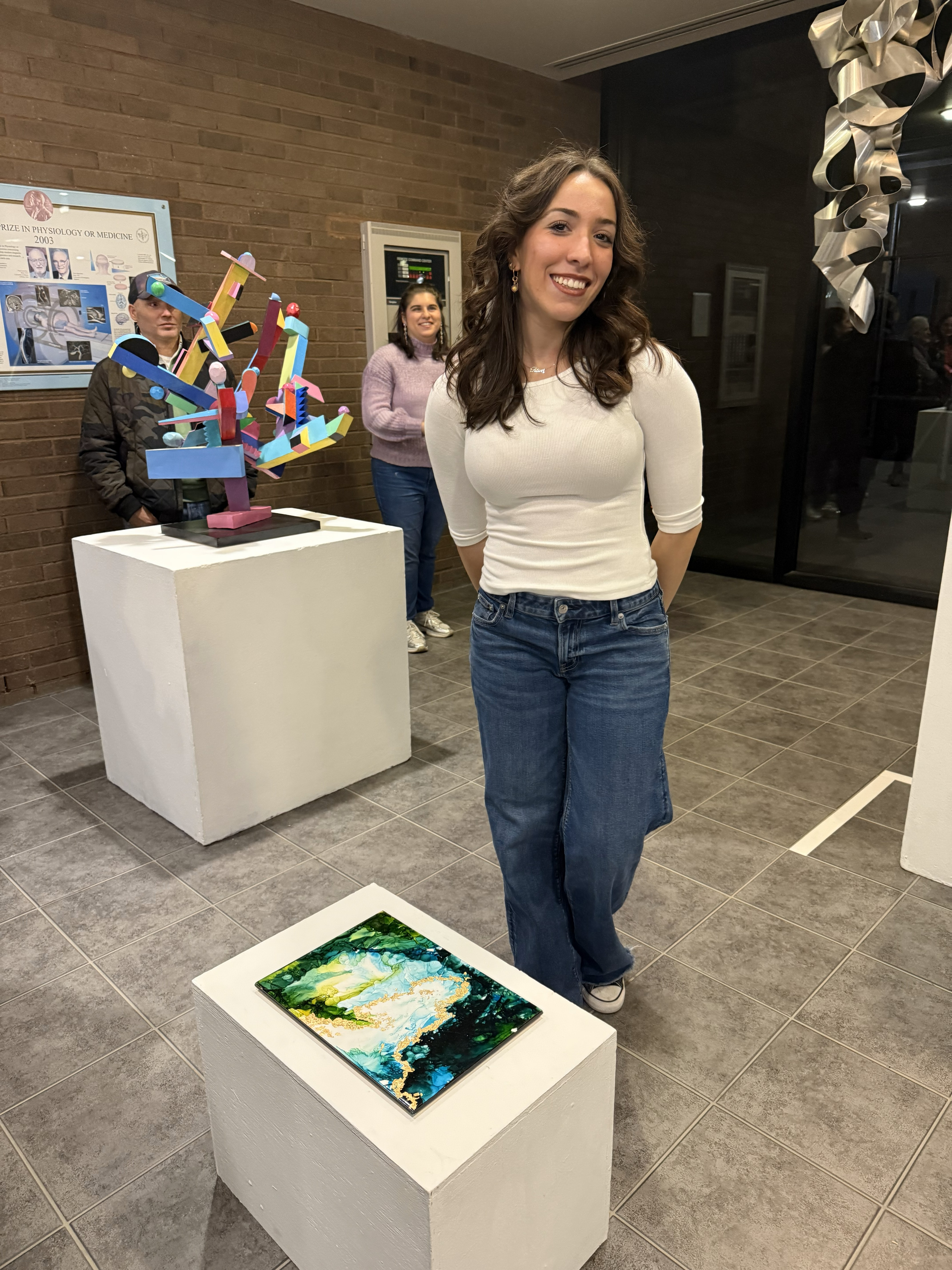 Eleni Tsevis Art Art student, Eleni Tsevis wearing a white shirt and jeans stands next to her green and gold acryclic art sculpture