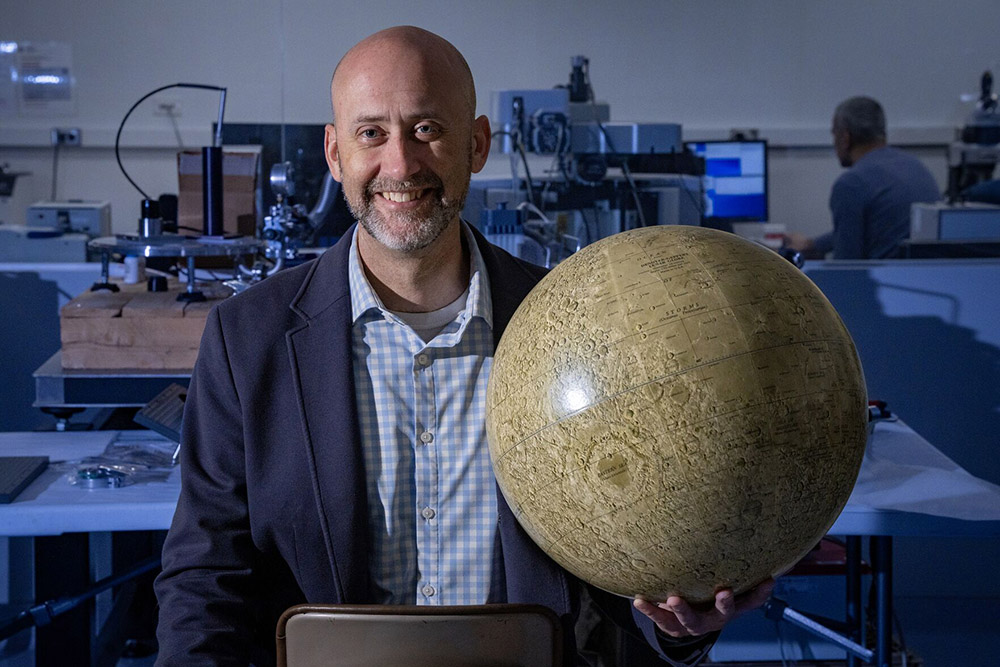 Timothy Glotch  wearing a blue and white checkered shirt and blue blazer  holds a moon model standing in a lab