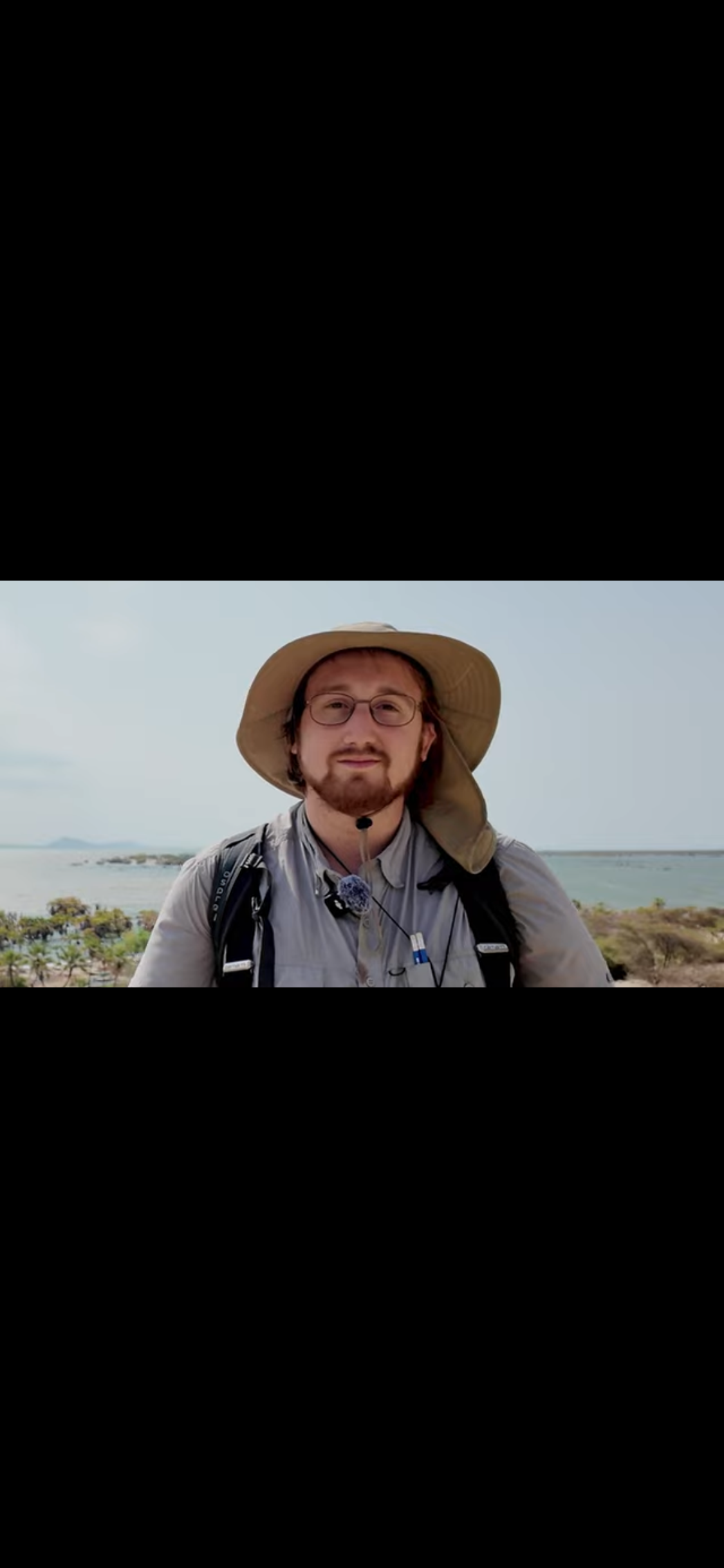 A person wearing a wide-brimmed hat and glasses stands outdoors with a backpack, looking at the camera. 
