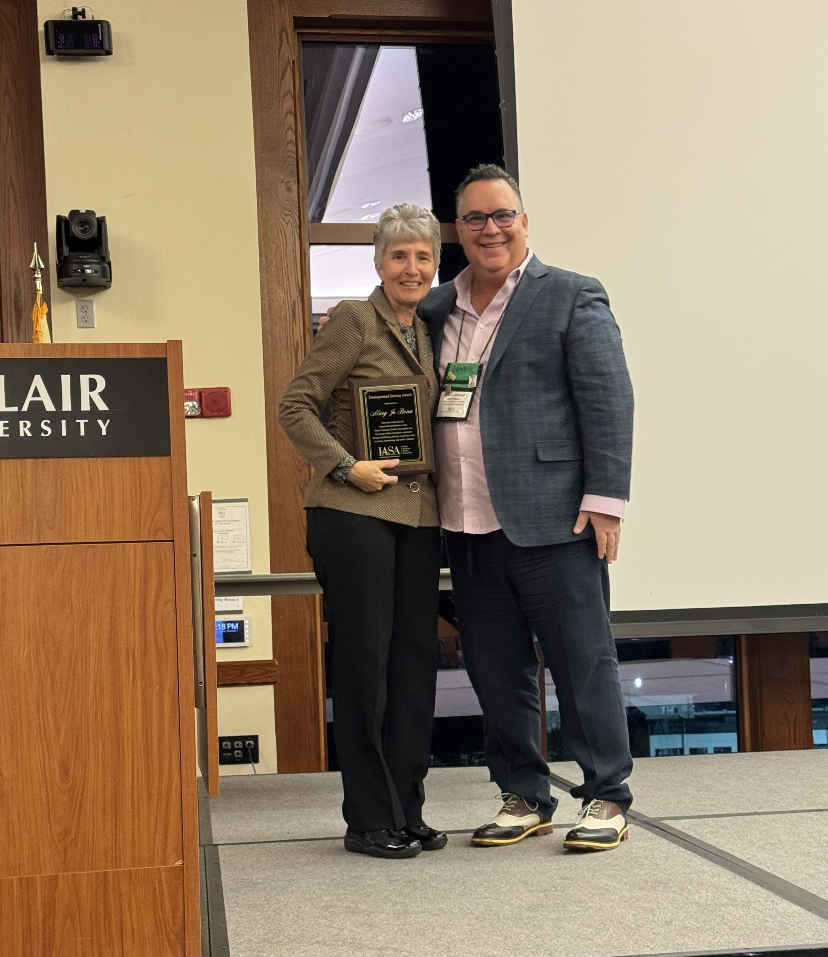 photo of Mary Jo Bona and Alan Gravano at the IASA conference. They're on a stage, by a podium, and Bona is holding her award