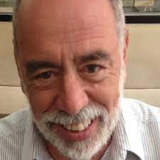 A professional, close-up headshot of a man with a warm and approachable expression. He has short grey hair and a well-groomed, full white beard and mustache. He is wearing a white and grey pinstriped dress shirt. The lighting is soft and even, highlighting his features against a neutral, out-of-focus background.