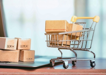 A miniature shopping cart beside a laptop with small shipping boxes strewn throughout