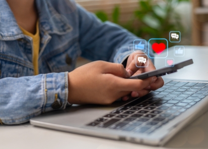 A pair of hands holding a phone over a laptop with social media like and comment icons floating overtop
