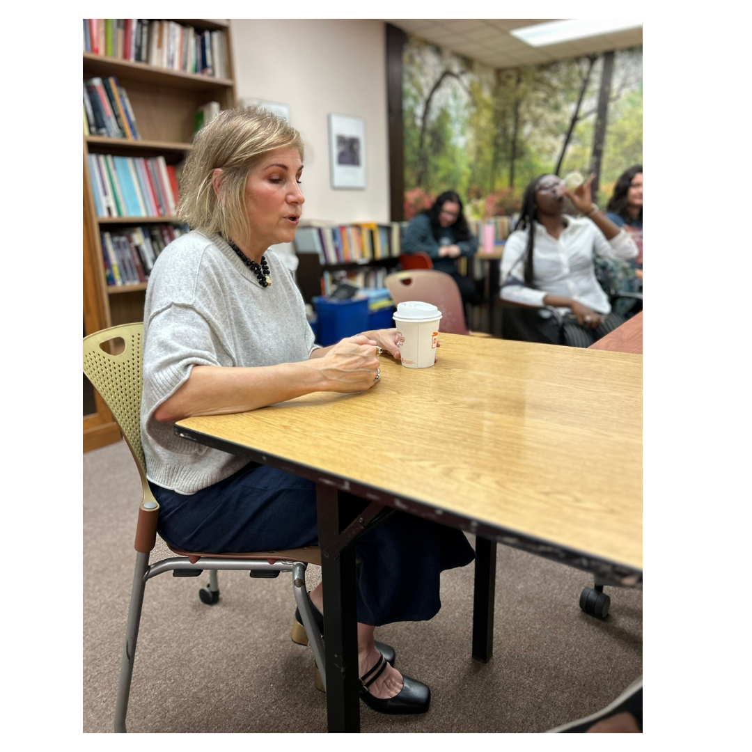 A woman sitting at a library table holding a paper cup, with bookshelves and a mural of trees in the background.