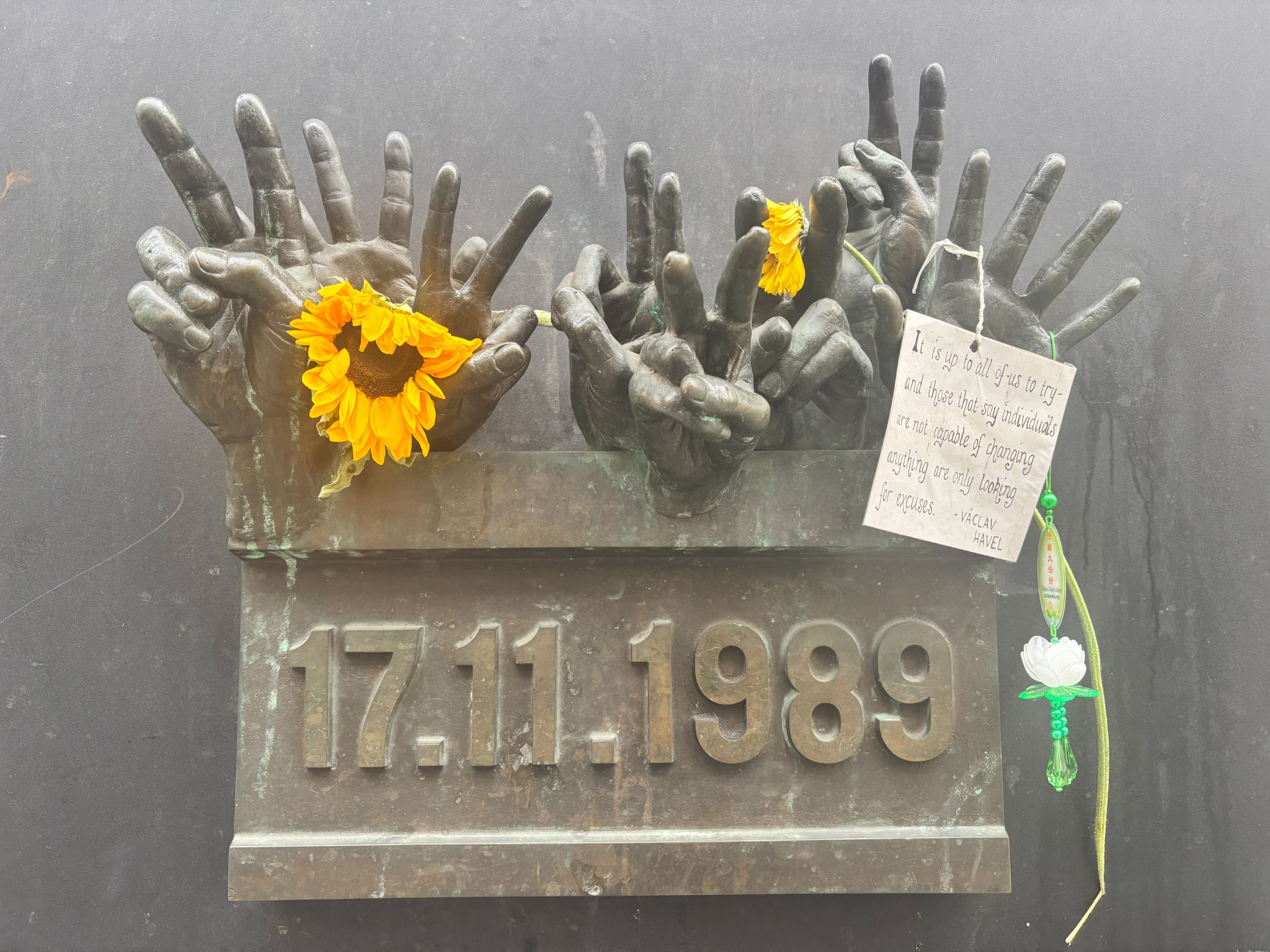 Czech Republic Democracy Statue Bronze sculpture of multiple hands reaching upwards, each adorned with yellow flowers and a green four-leaf clover. A plaque with the dates "17.11-1989" and a note with a message are also visible.