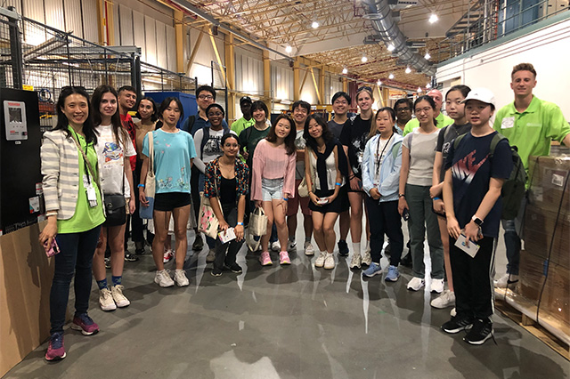 A group of 20 young adults and two guides in green shirts pose inside a warehouse. The atmosphere is cheerful and the setting is industrial.