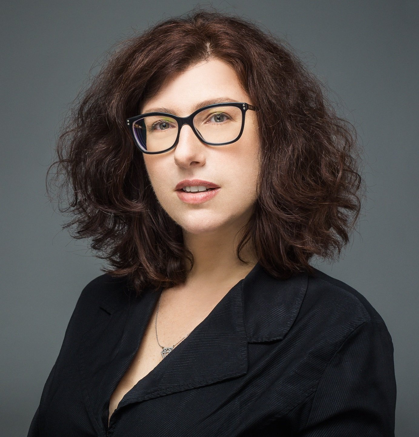 Pazit Ben-Nun Bloom A portrait of a woman with voluminous, shoulder-length curly dark brown hair and black-rimmed glasses. She is wearing a black blazer and looking toward the camera with a neutral expression. The background is a solid, dark gray.
